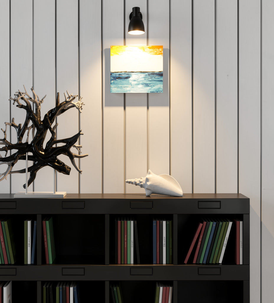 "Glow" is original abstract canvas wall art of a sunset sky over the ocean in orange, yellow, and blue, by Little Haven Art Studio, hanging in a coastal style room above a black bookcase with a shell.