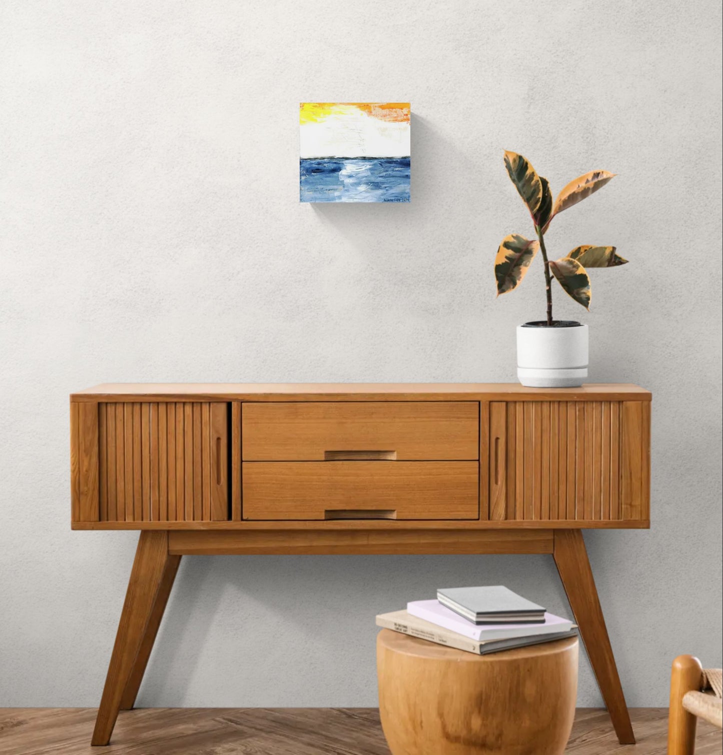 "Glow" is original abstract canvas wall art of a sunset sky over the ocean in orange, yellow, and blue, by Little Haven Art Studio, hanging above a wood cabinet with plant.