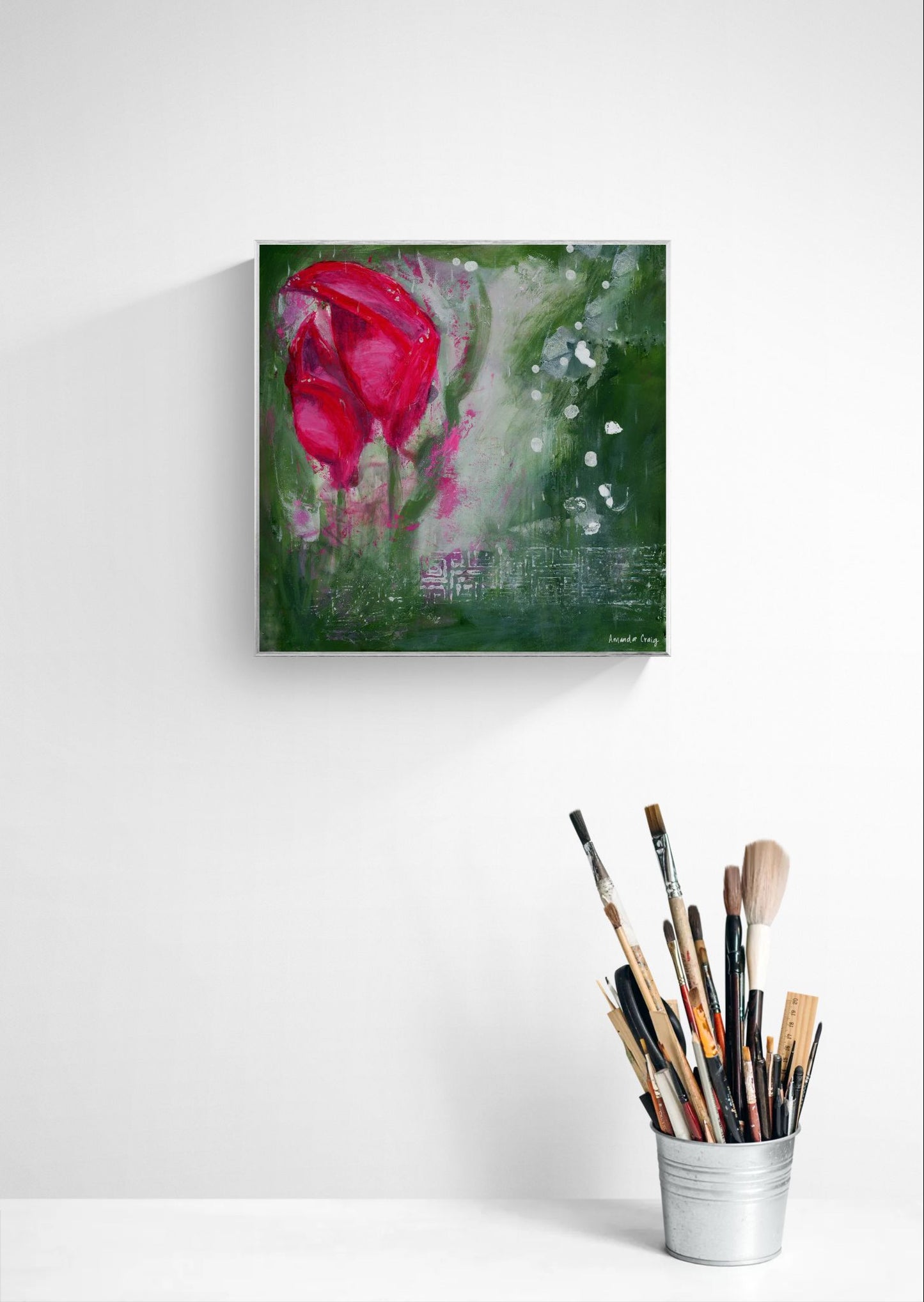 Original floral mixed media painting on abstract background titled, "Living Hope," by Amanda Craig of Little Haven Art Studio, in magenta pink, ruby red, emerald green, gray, and metallic silver, with silver tone floating frame hanging on a gray wall above paintbrushes.